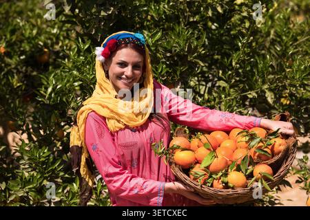 Gioiosa donna mediorientale con tradizionale abito rosa ricamato colorato e velo giallo con cesto in tessuto pieno di fresco raccolto Foto Stock