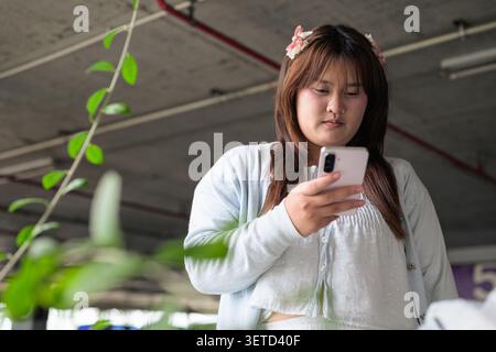 Un ritratto orizzontale di stile di vita di una adolescente Isan tailandese del sud-est asiatico concentrata sullo schermo del suo telefono. Ripresa da un angolo basso all'interno di un cov Foto Stock