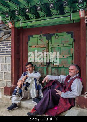 Due uomini in Hanbok maschile seduti davanti a un cancello verde in legno, da vicino ai tradizionali dettagli architettonici al Palazzo Gyeongbokgung di Seoul. Editoriale. Foto Stock