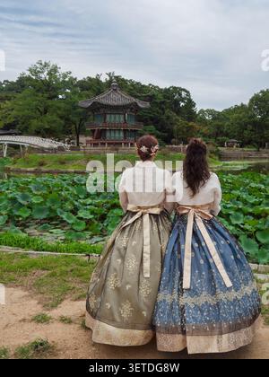 Vista posteriore di due giovani donne vestite con eleganti gonne tradizionali in hanbok coreano in piedi accanto a un laghetto di loto, godendosi la vista panoramica. Foto Stock
