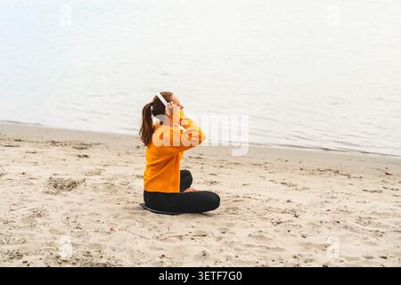 Donna rilassata che indossa le cuffie sedute sul fiume sulla sabbia e che si gode all'aperto. Foto Stock