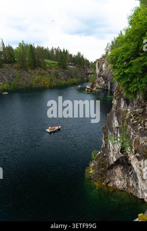 Ambiente tranquillo all'aperto con una piccola barca che trasporta persone in acque calme, circondata da aspre pareti di pietra, alberi sempreverdi e cielo nuvoloso luminoso Foto Stock