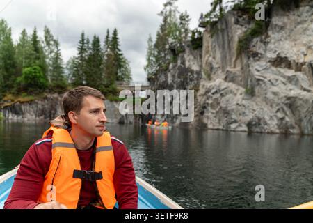 Il giovane con il giubbotto di salvataggio arancione si siede su una piccola barca sul tranquillo lago di montagna, con ripide coste rocciose, pini, cielo nuvoloso e acqua riflettente Foto Stock