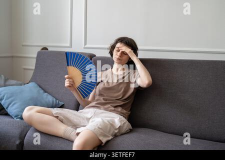 Donna seduta sul divano e rinfrescata, ventaglio di carta a mano, soffre di caldo estivo, faccia infastidita. Foto Stock
