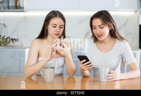 Due giovani donne che guardano il telefono e bevono tè in cucina Foto Stock