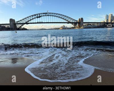 Un'onda bagna una spiaggia sabbiosa del porto di Sydney all'alba con il Sydney Harbour Bridge e l'Opera House visibili sull'acqua. Foto Stock