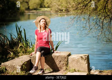 Donna con cappello di paglia e maglietta rossa seduta accanto a un lago tranquillo godendosi il sole Foto Stock