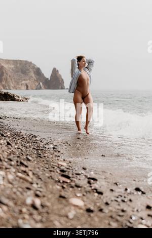 Donna vacanza al mare giovane donna in costume da bagno e camicia si erge sulla spiaggia di ciottoli con onde oceaniche e scogliere rocciose Foto Stock