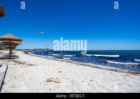Vista della Spiaggia sa Petra Ruja con ombrelloni di paglia su sabbia bianca, onde turchesi del mare e kitesurfer in lontananza sotto un cielo blu limpido a Sini Foto Stock