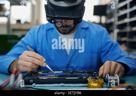 Tecnico addetto alla saldatura tecnico vestito con un camice da laboratorio blu sta riparando diligentemente un computer portatile Foto Stock