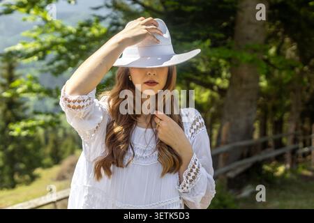 Donna in abito bianco che tiene il cappello nella foresta. Una donna con un abito in pizzo bianco regge un ampio cappello sul viso accanto a un sentiero nella foresta, con luce soffusa del sole Foto Stock