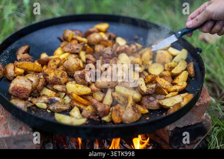 Patate e carne fritte sul fuoco all'aperto. Patate e carne friggono in una padella nera su un fuoco aperto, con fumo, fiamme e una mano che tiene un coltello Foto Stock