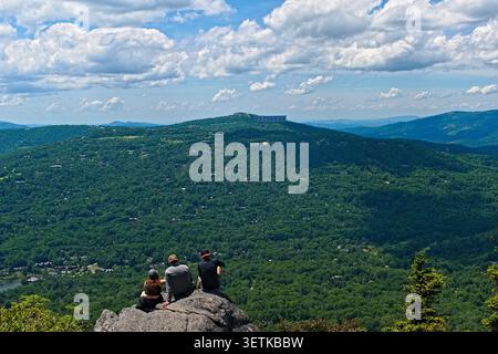 Un gruppo di persone si siede su una sporgenza rocciosa, osservando un'ampia vista di lussureggianti montagne verdi e lontane colline blu sotto un cielo nuvoloso. Foto Stock