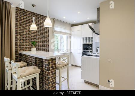 Cucina moderna caratterizzata da una parete d'accento in mattoni con un controsoffitto bianco isola e sgabelli da bar. Armadi e elettrodomestici bianchi completano il look, con Pend Foto Stock