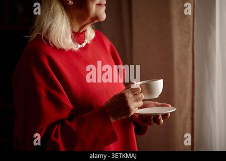 Donna anziana che tiene una tazza di caffè vicino alla finestra in mattinata tranquilla Foto Stock