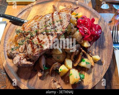 Primo piano ad angolo alto che mostra bistecca di carne grigliata con timo, servita con patate arrosto, funghi e cipolla rossa sottaceto su tavola rotonda di legno Foto Stock