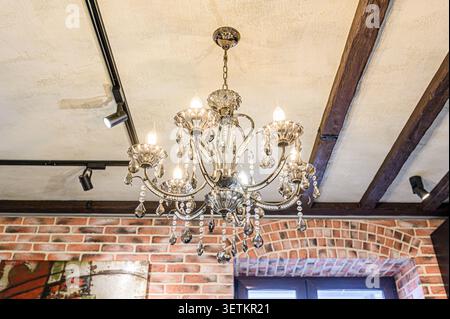 Un lampadario di cristallo è appeso sotto un soffitto con travi in legno a vista e illuminazione a binario. La muratura è visibile nella parte inferiore del telaio Foto Stock