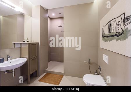 Bagno moderno con piastrelle beige neutre, lavandino e WC bianchi, cabina doccia e un'estetica minimalista. Un disegno di esopo incorniciato aggiunge un a. Foto Stock