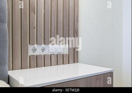 Primo piano di un interno moderno con parete inclinata in legno, prese elettriche e interruttori luminosi incorporati sopra un ripiano bianco Foto Stock