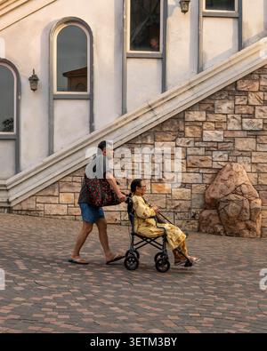 13.12.25 Sunset Town, isola di Phu Quoc, Vietnam. Strade prima dell'anno nuovo. Un uomo spinge una donna anziana su una sedia a rotelle lungo un sentiero lastricato in una città turistica. Foto Stock