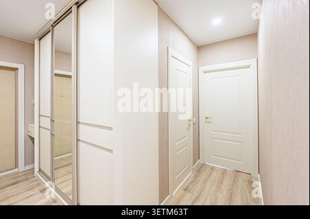 Un corridoio ben illuminato con un grande armadio bianco con porte a specchio, due porte bianche e pavimento in legno. Le pareti neutre completano la scena Foto Stock