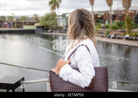 Una donna si trova davanti a un tranquillo lungomare, osservando attentamente le acque calme. Porta con sé una borsa elegante, vestita con una giacca bianca, persa in riflessi Foto Stock