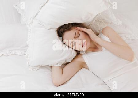 Una giovane donna giace a letto, visibilmente dolorosa, tenendo la testa a causa di un mal di testa, circondata da morbidi cuscini bianchi, che illustrano i problemi di salute delle donne. Foto Stock