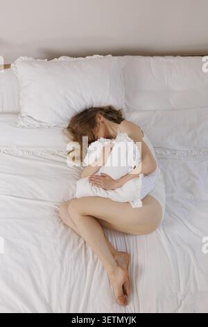 Una donna sta a letto, tenendo un cuscino da vicino mentre soffre dolore addominale durante il ciclo mestruale, evidenziando le sfide dell'hea femminile Foto Stock