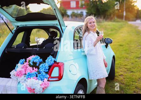 fuga in campagna caratterizzata da una donna che si rilassa accanto a un hatchback pastello decorato con ortensie nella calda luce serale Foto Stock