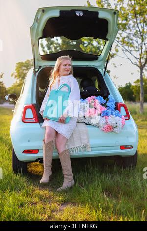 concetto di viaggio su strada con una giovane donna sorridente, veicolo pastello, ortensie fresche e luce dorata del sole serale in un campo tranquillo Foto Stock