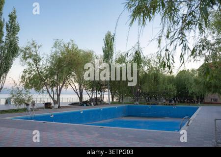 Ampia piscina all'aperto vuota con piastrelle blu e corrimano in metallo, circondata da un'area di mattoni pavimentati, con una fila di alberi verdi e una calma Foto Stock