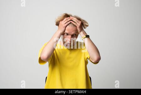 Giovane che afferra la testa con entrambe le mani, sperimentando stress e shock intensi, guardando lontano con incredulità, indossando una t-shirt gialla contro una Foto Stock