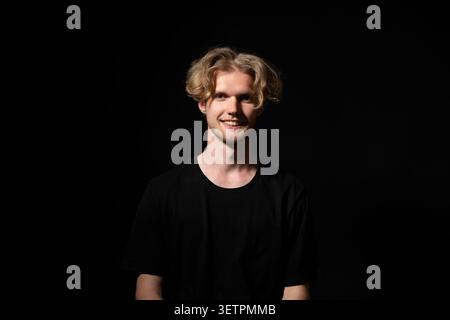 Giovane biondo sorridente, che esprime felicità e fiducia in un ritratto in studio, guardando direttamente la fotocamera su uno sfondo nero e neutro Foto Stock