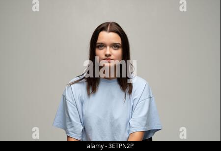 Giovane donna dall'espressione seria che indossa una t-shirt blu, guarda direttamente la fotocamera, ritratto in studio Foto Stock