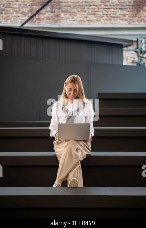 Donna di mezza età sicura e concentrata che lavora con un notebook mentre è seduta all'aperto su una moderna scalinata urbana. Foto Stock