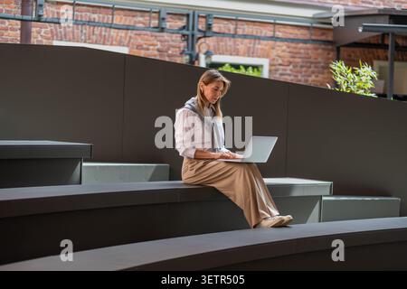 Focalizzata sul laptop di una donna di mezza età, freelance lavora all'aperto, siede su una moderna panchina in un luogo pubblico. Foto Stock