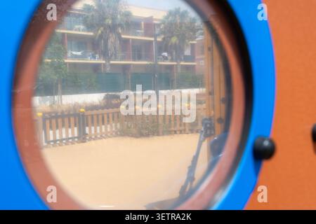 Una prospettiva ravvicinata attraverso un'apertura circolare rivela un'area giochi soleggiata con una recinzione in legno e un edificio moderno con balconi e palme Foto Stock