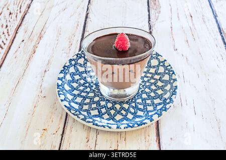 Una crema di mousse al cioccolato ricoperta da un lampone fresco si trova in una tazza di vetro su un piatto decorativo bianco e blu. Foto Stock