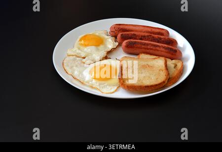 Colazione, due uova fritte, salsicce e pane tostato in un piatto rotondo su sfondo nero. Primo piano. Foto Stock