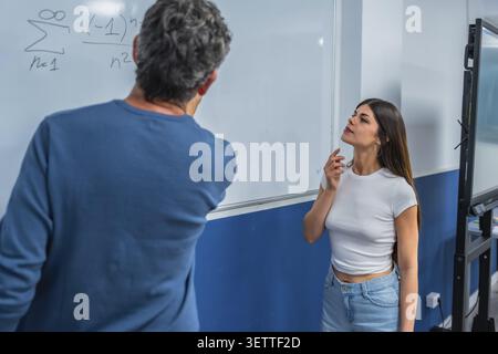 L'istruttore scrive complesse equazioni matematiche su una lavagna, spiegando il problema a una giovane studentessa attenta che impara e capisce Foto Stock