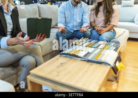 Una giovane coppia si consulta con un assistente di vendita in uno showroom di mobili, confrontando campioni di tessuto e colori per scegliere il divano perfetto per la loro nuova struttura Foto Stock