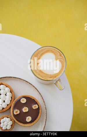 Latte sospeso con schiuma di cuore accanto alle crostate da dessert su un tavolo bianco, sfondo giallo Foto Stock