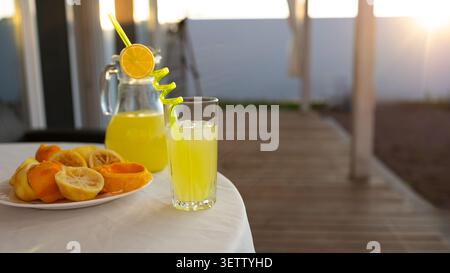 Limonata di colore giallo brillante servita in vetro trasparente con paglia riccia e ruota di limone, caraffa corrispondente ripiena di agrumi e piatto di limoni affettati Foto Stock