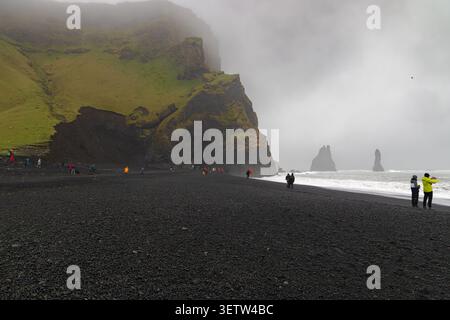 Turisti che camminano sulla spiaggia di sabbia nera di Reynisfjara in Islanda vicino a scogliere di basalto, scogliere marine e onde oceaniche sotto un cielo nebbioso. Foto Stock
