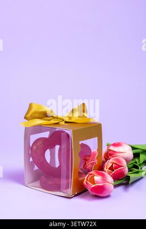 Un'ampia foto pubblicitaria con una confezione regalo di souvenir dorata legata a un nastro di raso e un piccolo bouquet di tulipani rosa freschi su una vivace ba di lavanda Foto Stock