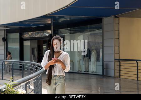 giovane donna con lunghe trecce scure, indossa una blusa in pizzo bianco e jeans, in piedi in un centro commerciale aperto Foto Stock
