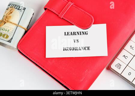 Un taccuino rosso si trova su un tavolo accanto a una pila di soldi e a una calcolatrice. Sul notebook viene posizionata una scheda con le parole apprendimento e ignoranza. Foto Stock