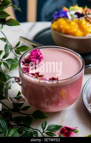 Una tazza di vetro riempita con un latte matcha rosa schiumoso ricoperto di petali secchi e un vivace fiore rosa, con un recipiente per frullati sfocato sullo sfondo. Foto Stock