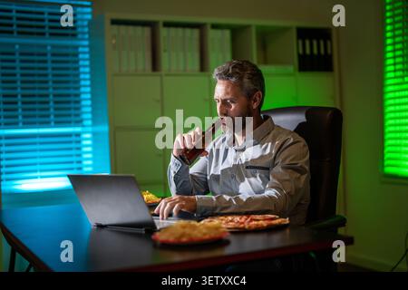 Uomo d'affari che lavora fino a tardi per guardare contenuti online. Libero professionista che lavora fino a tardi con un umore rilassato. Uomo ispanico anni '40 che guarda un portatile. Il freelance mangia la pizza Foto Stock