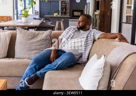 Uomo sdraiato sul divano componibile beige in un'area soggiorno open space, con camicia a righe e jeans Foto Stock